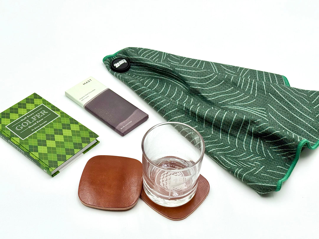 Luxury golf gift set with green magnetic golf towel, whiskey glass, leather coasters, golfer book, and dark chocolate bar.
