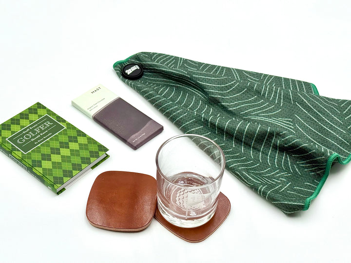 Luxury golf gift set with green magnetic golf towel, whiskey glass, leather coasters, golfer book, and dark chocolate bar.