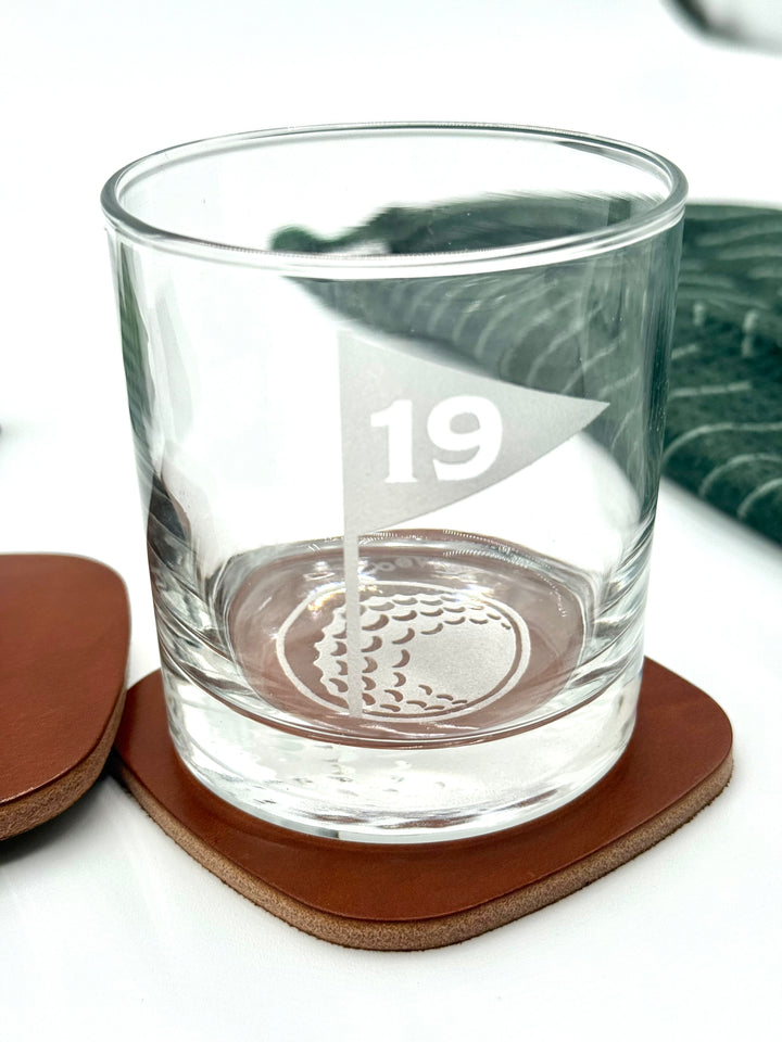 Whiskey rocks glass etched with 19th hole flag and golf ball, displayed on premium leather coaster.