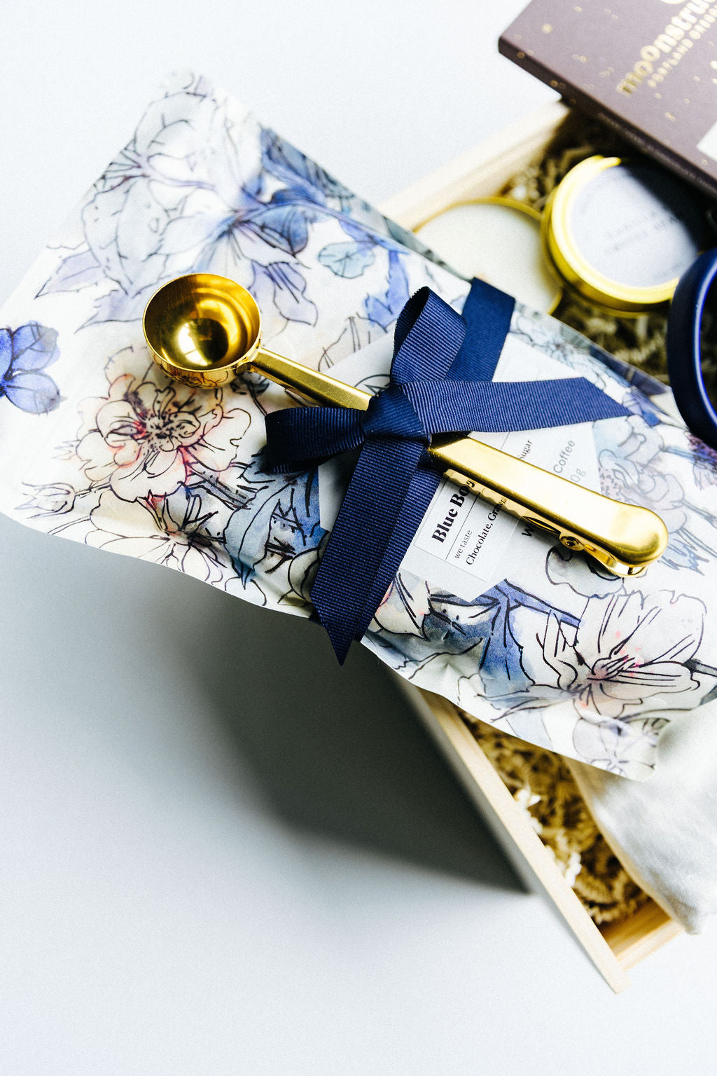 Luxury coffee gift box close-up featuring Methodical Blue Boy Coffee wrapped in floral packaging, tied with navy ribbon, and gold coffee scoop clip.