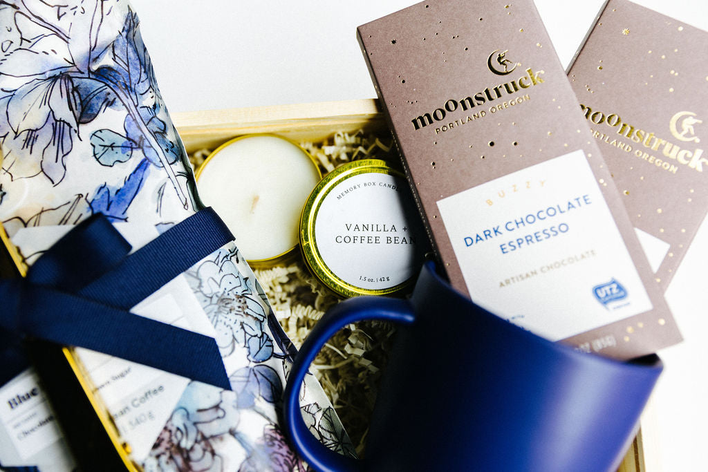 Grounded gift box contents with navy ceramic mug, vanilla and coffee bean soy candle, and two Moonstruck dark espresso chocolate bars.