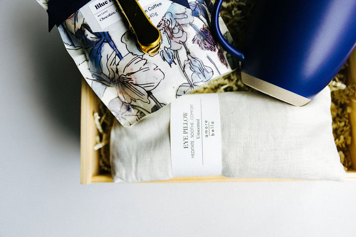 Ivory linen unscented eye pillow and navy mug resting beside floral coffee packaging in wooden gift box.