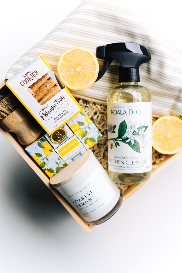 Close-up of Coastal Lemon candle, lemon soap, eco-friendly kitchen cleaner, wooden scrub brush, and taupe-striped ruffle towel for housewarming gift box