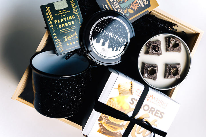 Overhead view of open wooden box with fire pit, mug, sticker, and playing cards arranged with black crinkle filler