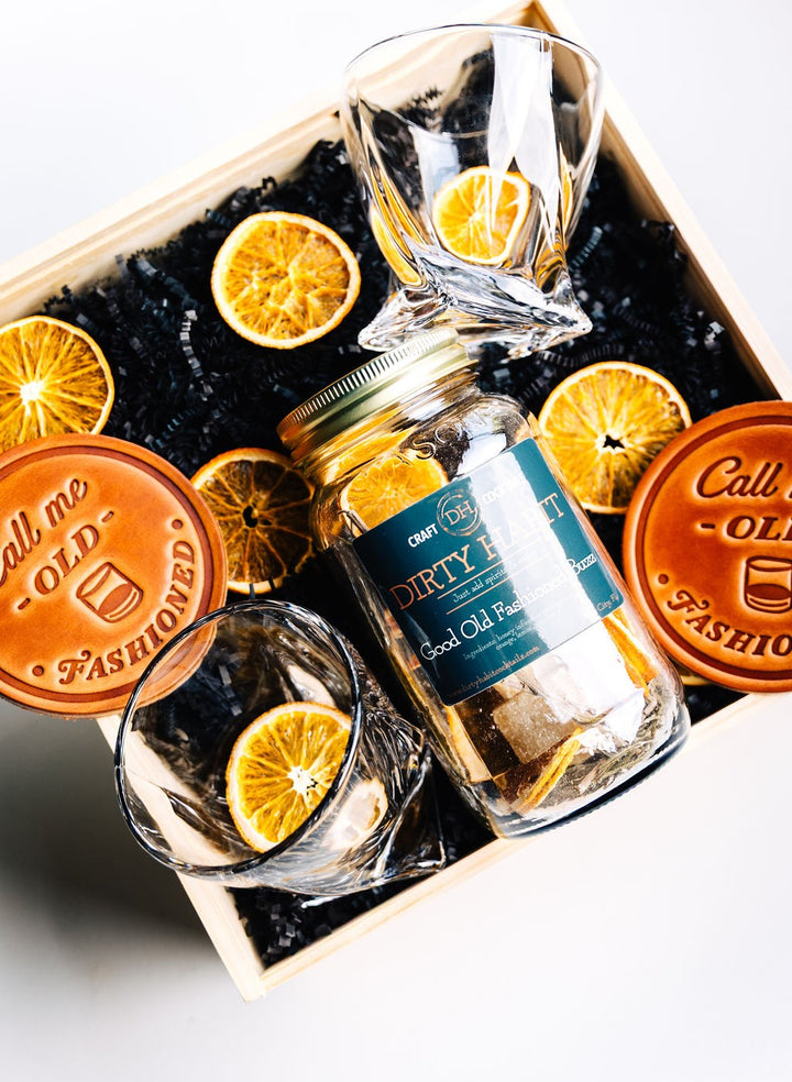 Flat lay of whiskey glasses on leather coasters with Dirty Habit cocktail mix, dried orange slices, rosemary sprigs, and sugar cubes