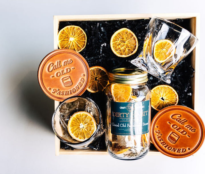 Luxury Old-Fashioned gift box with cocktail mix jar, crystal twist whiskey glasses, leather coasters, and dried orange garnish in wooden keepsake box