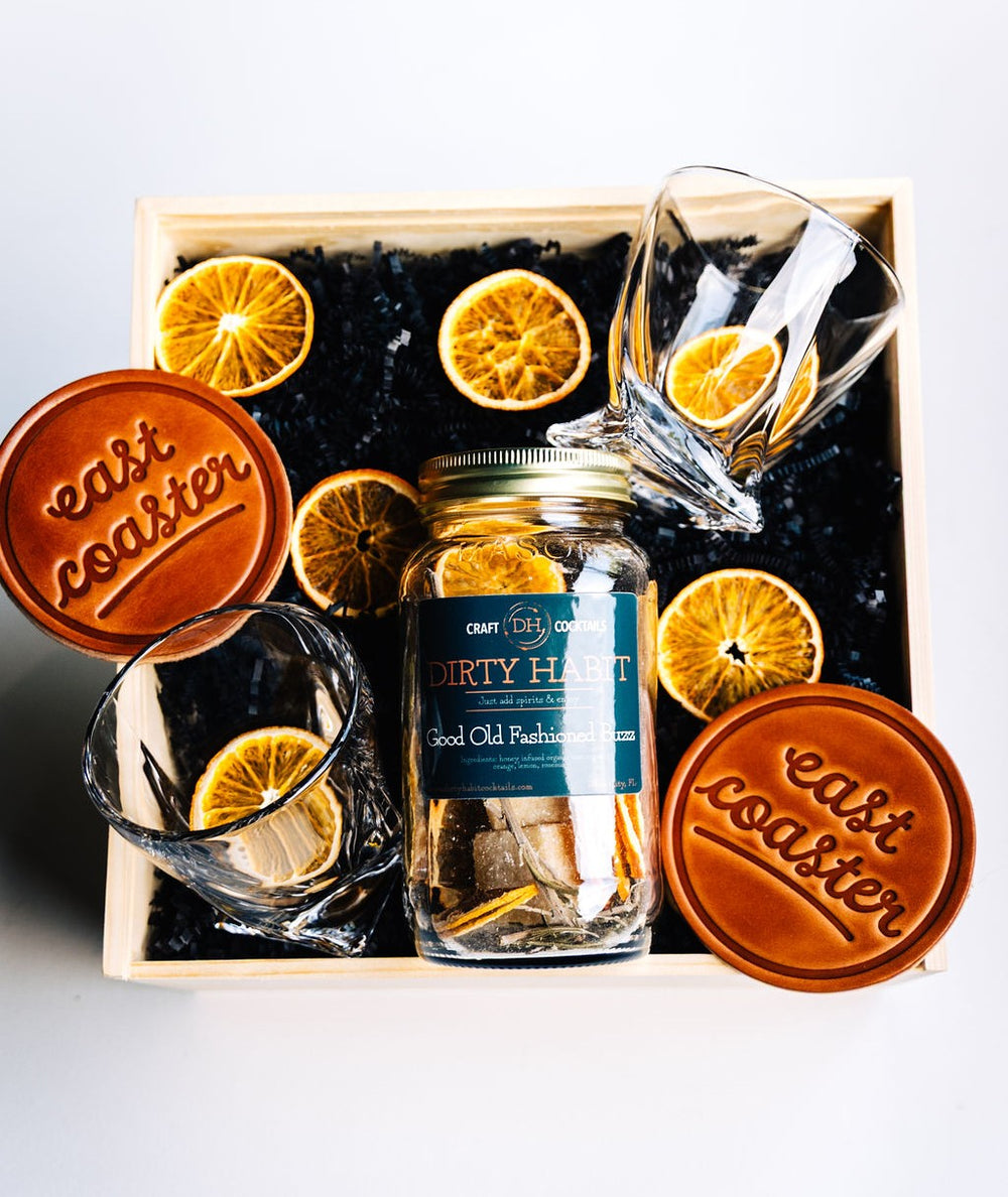 Top-down view of The Old-Fashioned Way gift box featuring cocktail mix, two crystal whiskey glasses, "east coaster" leather coasters, and dried oranges