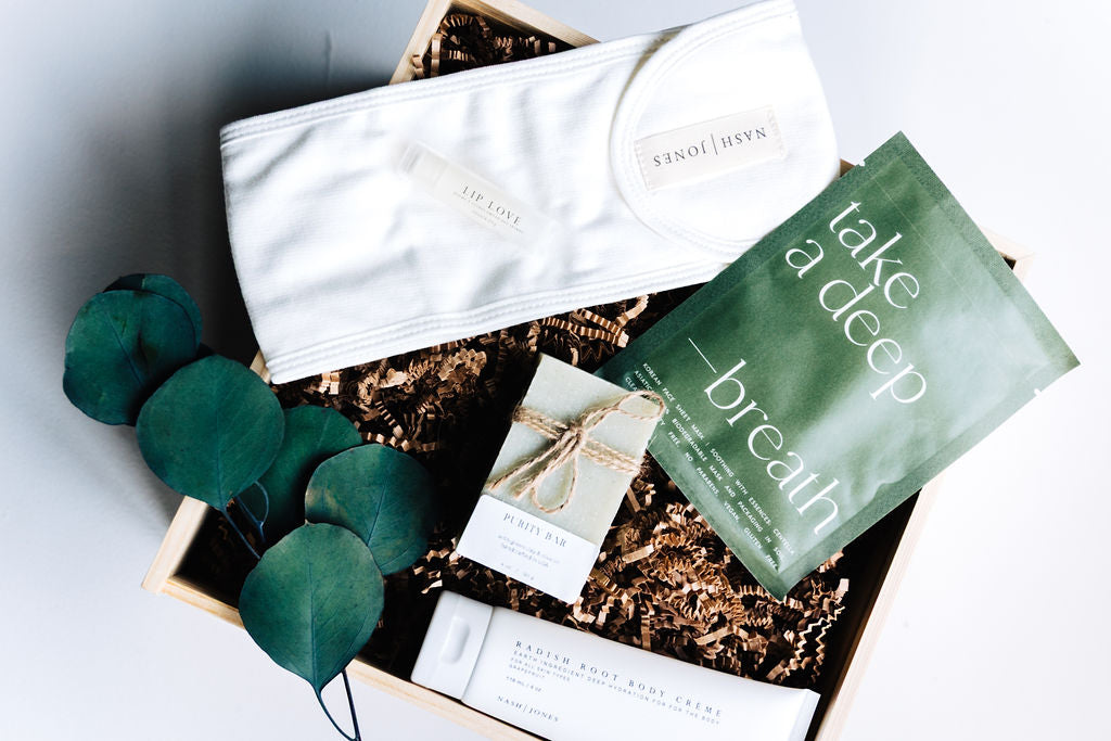 Luxury wooden gift box with bamboo spa headband, biodegradable sheet mask, organic soap bar, body crème, and lip balm arranged with natural filler.