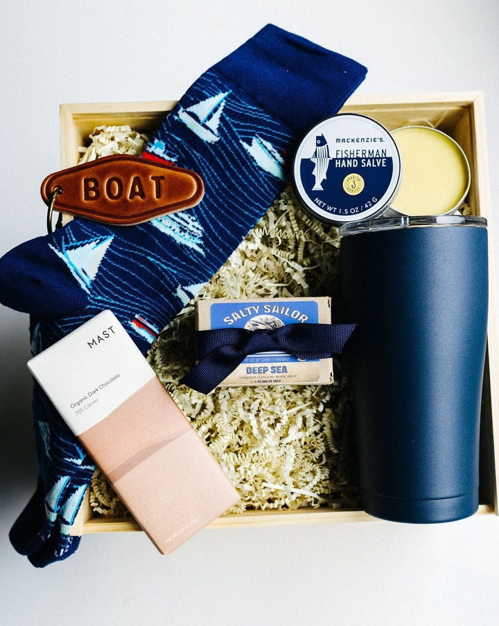 Overhead view of nautical Salty Sailor Gift Box with navy tumbler, yacht-patterned socks, leather keychain, bar soap, chocolate, and hand salve