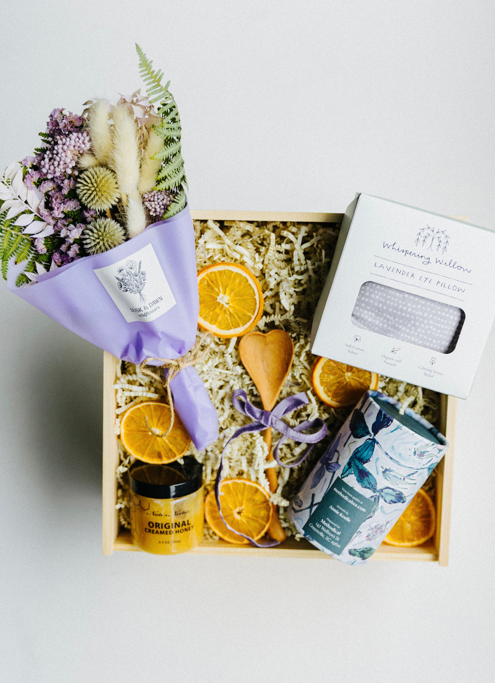 Top view of A Gentle Pause relaxation gift box featuring tea, honey, lavender eye pillow, wooden spoon, and violet dried flower bouquet.
