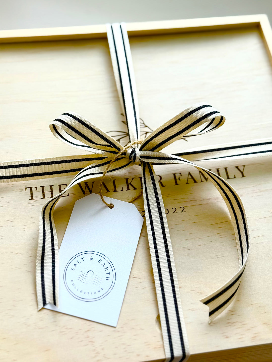 Personalized wooden gift box lid engraved with family name, tied with striped ribbon and Salt & Earth gift tag.