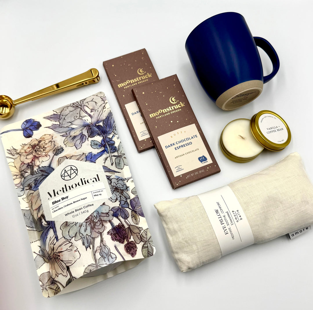 Flat lay of Grounded Coffee Lover’s Gift Box contents – Methodical Blue Boy Coffee in floral wrap with gold coffee scoop clip, navy ceramic mug, vanilla and coffee bean soy candle in gold tin, ivory linen eye pillow, and two Moonstruck dark espresso chocolate bars.