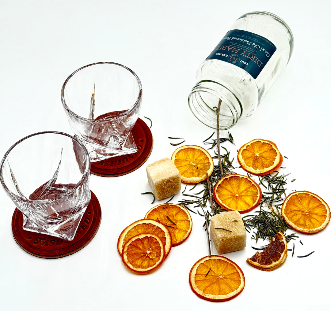 Flat lay of whiskey glasses on leather coasters with Dirty Habit cocktail mix, dried orange slices, rosemary sprigs, and sugar cubes