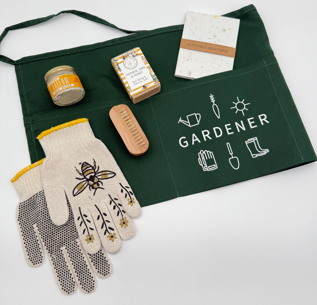 Gardener apron, bee gloves, seed paper notebook, natural soap, nail brush, and tallow body butter arranged on white background