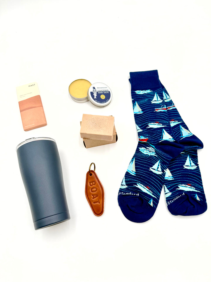 Flat lay of Salty Sailor Gift Box items – navy tumbler, yacht socks, leather BOAT keychain, hand salve, bar soap, and Mast dark chocolate