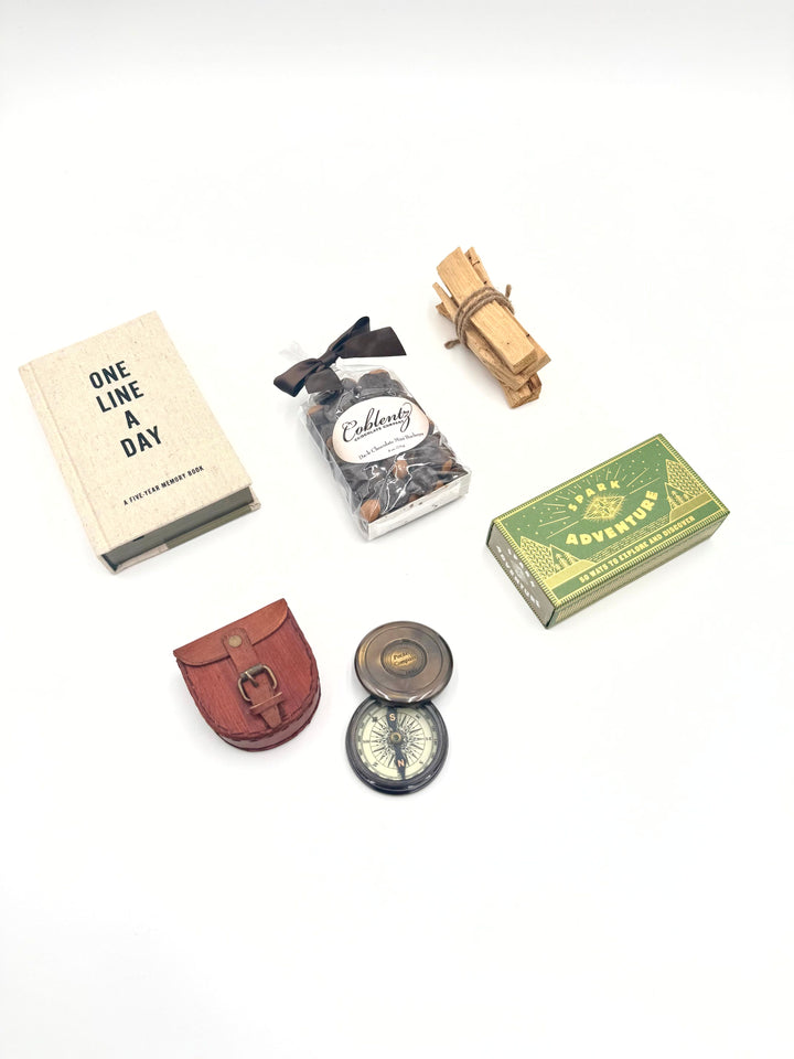 Adventure Awaits gift box contents displayed flat lay: compass with leather case, Spark Adventure box, journal, Palo Santo sticks, and chocolate buckeyes
