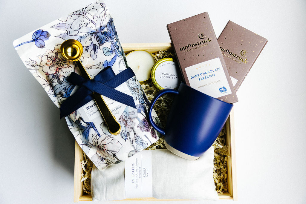 Luxury coffee gift box with artisan coffee beans, navy ceramic mug, vanilla candle, eye pillow, and dark chocolate bars.