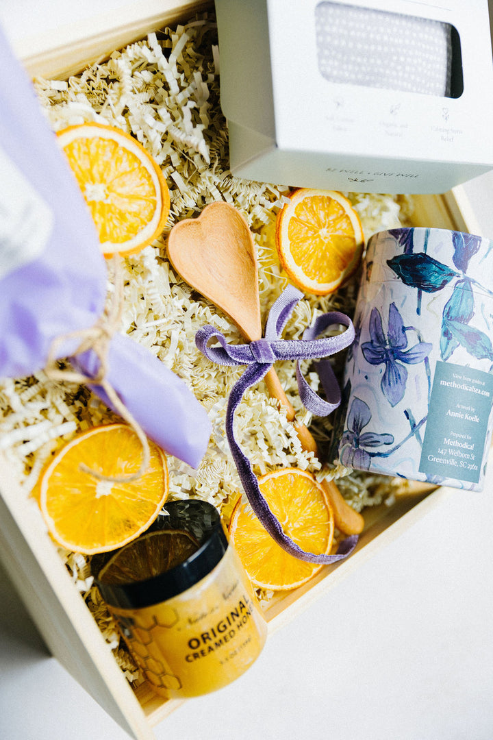 Flat lay of A Gentle Pause gift box with Earl Grey tea tin, creamed honey, lavender eye pillow, heart-shaped spoon, and dried citrus accents.