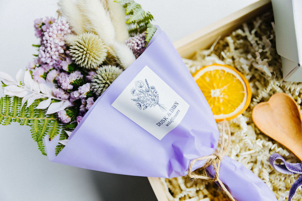 Mini bouquet of dried violet blooms wrapped in lavender paper, placed inside luxury wooden gift box with heart-shaped spoon and dried orange slices.